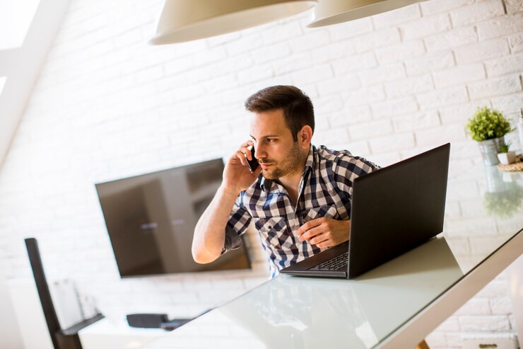Foto joven sentado en el escritorio de la cocina en casa y usando una computadora portátil y un teléfono móvil