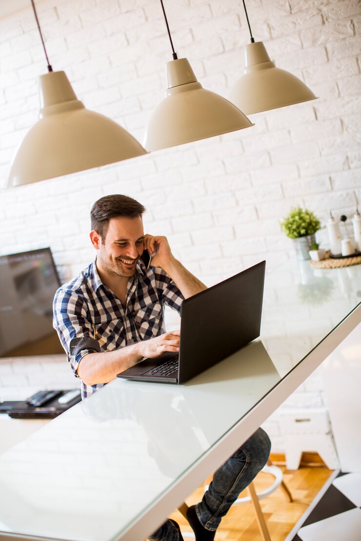 Foto joven sentado en el escritorio de la cocina en casa y usando una computadora portátil y un teléfono móvil