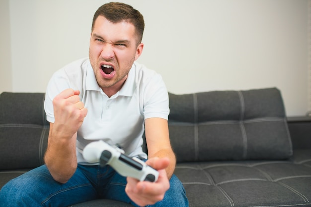 Joven sentado en la habitación jugando juegos de computadora. Persona de sexo masculino mantenga joystick en la mano y animar con placer. Gritando y gritando durante el juego.