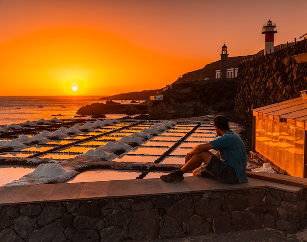 Un joven sentado en la puesta de sol del Faro de Fuencaliente junto a la mina de sal, en la ruta de los volcanes al sur de la isla de La Palma, Islas Canarias, España