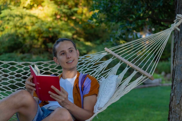 Foto joven sentado relajado en una hamaca leyendo un libro rojo y mirando hacia un lado disfrutando del entorno natural