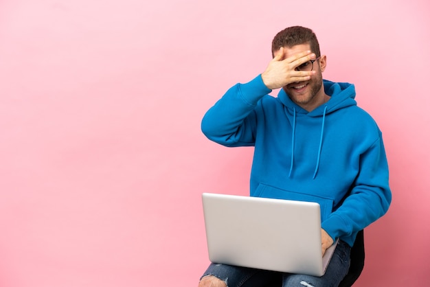 Joven sentado en una silla con laptop cubriendo los ojos con las manos y sonriendo