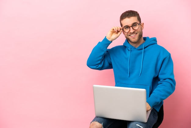 Joven sentado en una silla con laptop con gafas y feliz