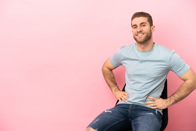 Joven sentado en una silla sobre fondo rosa aislado posando con los brazos en la cadera y sonriendo