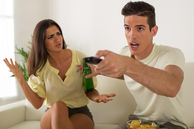 Joven sentado en el sofá y viendo la televisión. Su esposa enojada gritando.