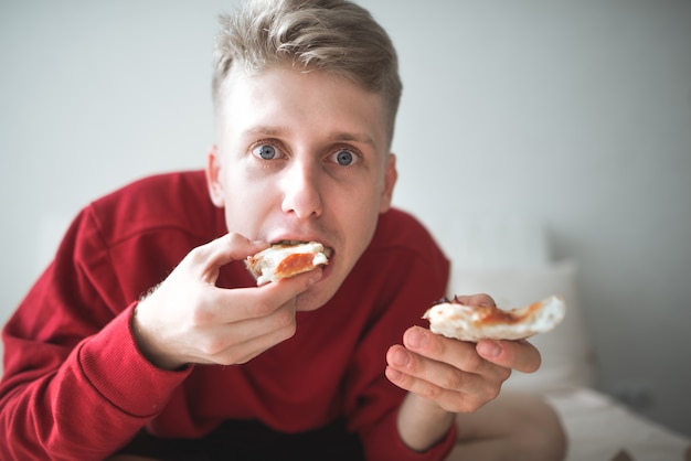 Joven sentado en su casa en la cama en una acogedora habitación comiendo pizza