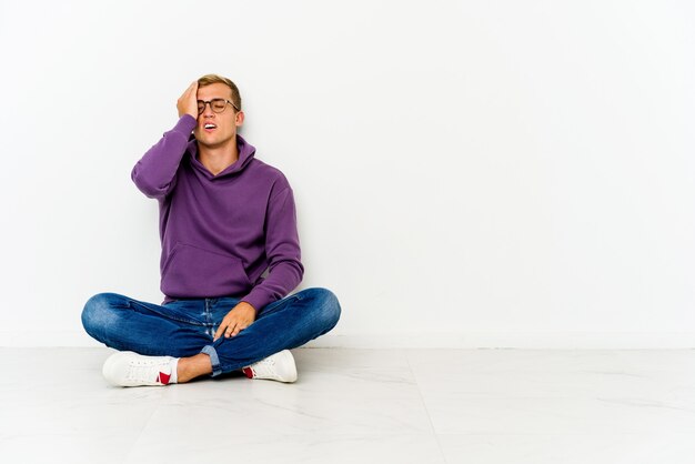 Joven sentado en el suelo olvidando algo, golpeándose la frente con la palma y cerrando los ojos