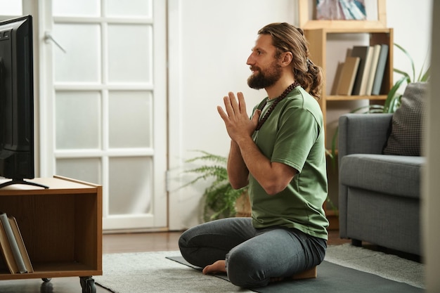 Joven sentado en el suelo en posición de loto y viendo un programa de yoga en la televisión en casa