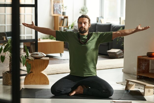 Joven sentado en un tapete de ejercicio con los ojos cerrados y disfrutando de ejercicios de yoga por la mañana en casa