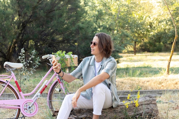 Joven sentado en el tronco de un árbol enviando mensajes de texto a sus amigos en el teléfono inteligente con una bicicleta vintage rosa