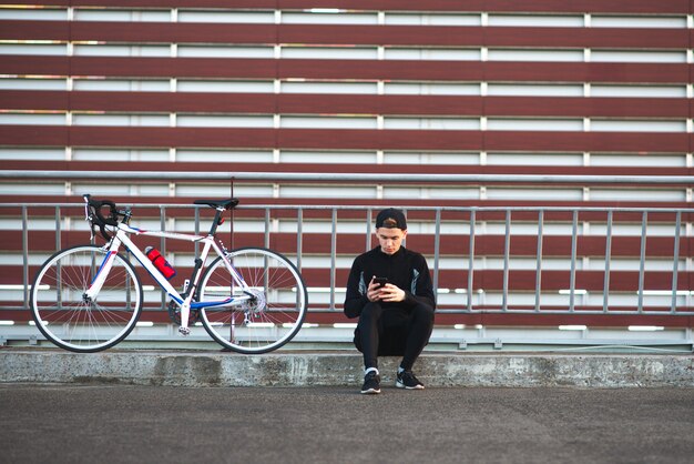 Joven sentado en un vestido oscuro con una bicicleta y usando un teléfono inteligente en el fondo de una pared de borgoña a rayas