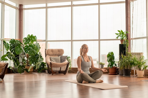 Joven serena mujer rubia en ropa deportiva cruzando las piernas mientras está sentado en la estera en pose de loto y practicando ejercicios de meditación en casa