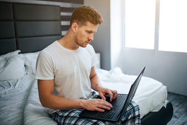 Joven serio en la cama esta mañana. El trabaja en casa. Escriba el tipo en el teclado del portátil y mire la pantalla. Luz.