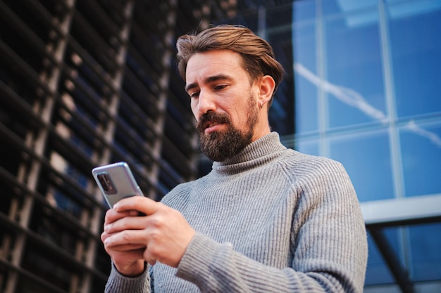 Un joven serio concentrado y preocupado usando una aplicación de teléfono móvil enviando mensajes de texto y compartiendo medios en un espacio de trabajo