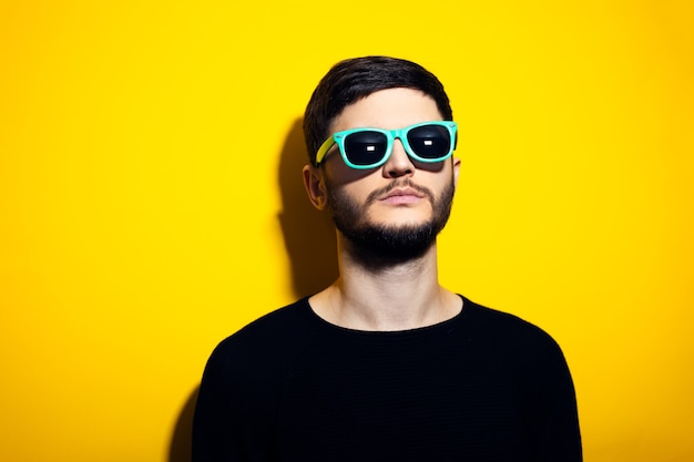 joven serio con gafas de sol cian y suéter negro en la pared amarilla.