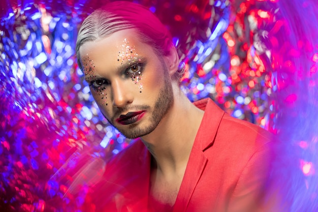 Joven serio con maquillaje escénico, cabello blanco con peinado preciso y elegante chaqueta roja sobre el cuerpo desnudo mirándote sobre fondo de colores