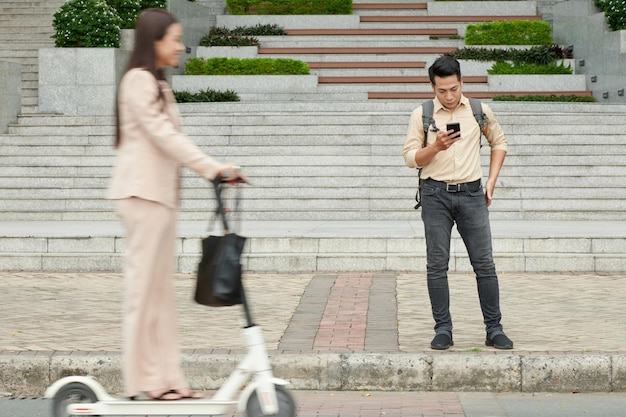 Un joven serio parado en el pavimento y leyendo un mensaje de texto en un smartphone cuando una mujer va en scooter