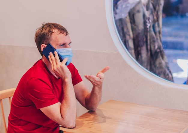 Joven serio, pensativo, atento, medio en mascarilla médica, pensamiento tenso, hablar, contestar el teléfono, en camiseta roja, en el café. La gente real expresa problemas de vida, resolviendo la comunicación durante la pandemia de Covid.