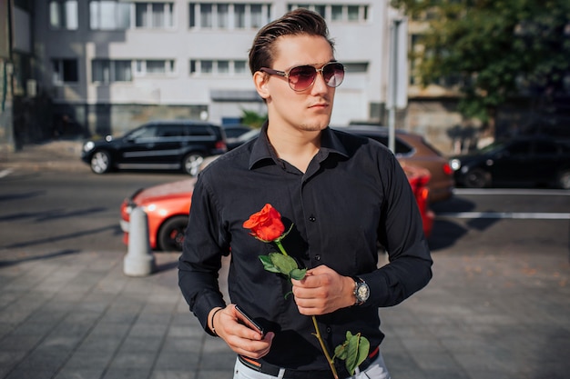 Joven serio usa gafas de sol y mira a la derecha. Él sostiene la rosa roja y el teléfono en las manos. El espera Está soleado afuera.