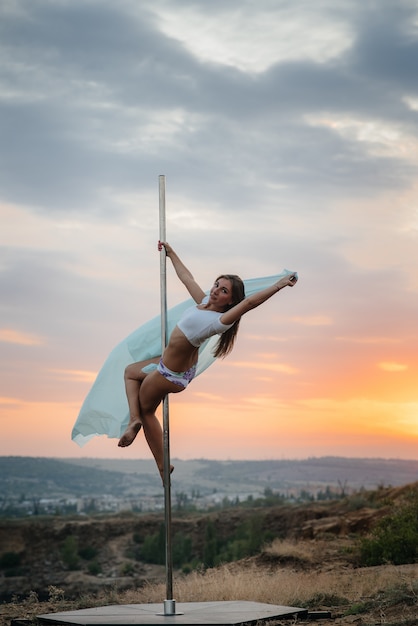 Una joven sexy realiza ejercicios increíbles en poste durante una puesta de sol. Baile. Sexualidad.