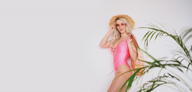 Joven sexy sonriendo feliz rubia en un traje de baño rosa, un sombrero de paja, gafas de sol, entusiasmados por presentar el producto. Mujer sobre un fondo blanco con hojas de palma verde.