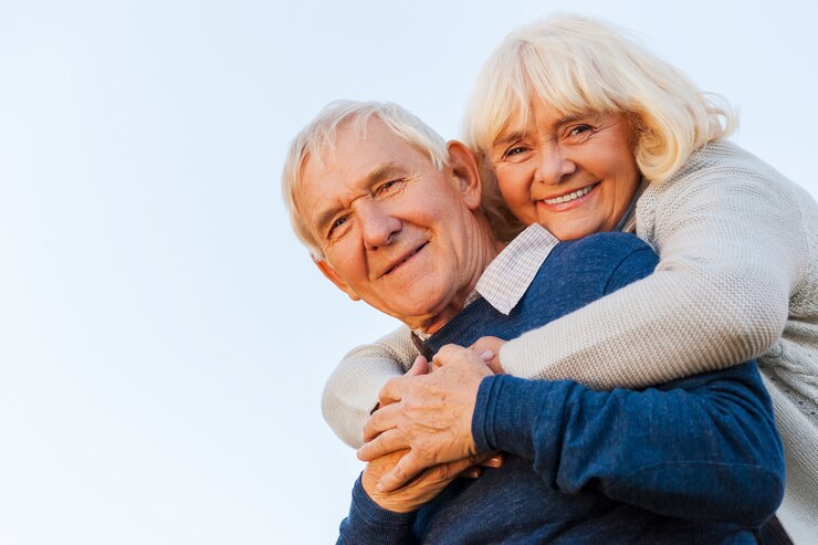 Joven para siempre. Vista de ángulo bajo de la feliz pareja senior uniéndose entre sí y sonriendo con el cielo azul como fondo