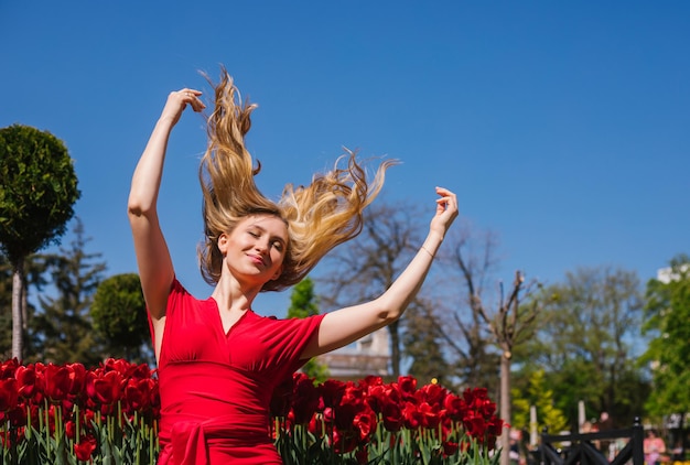 Una joven se sienta al sol entre los tulipanes Con un vestido rojo, su cabello ondeando al viento