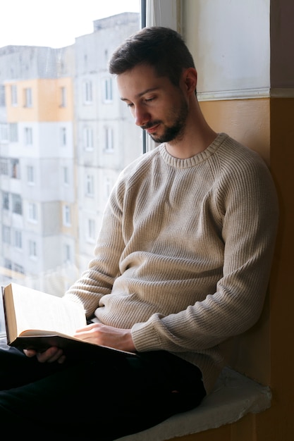 Un joven se sienta en el alféizar de una ventana y lee un libro. Un hombre lleva un suéter beige. El hombre tiene barba.