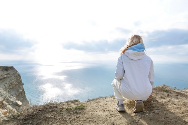 Una joven se sienta en una alta montaña y mira el mar y un hermoso paisaje en un día soleado chica rubia con el cabello que fluye en una chaqueta blanca estilo de vida activo y recreación vista trasera