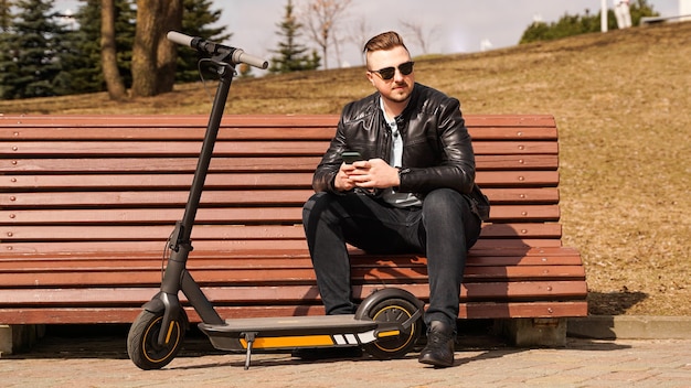 Joven se sienta en un banco en un scooter eléctrico de primavera junto a un banco