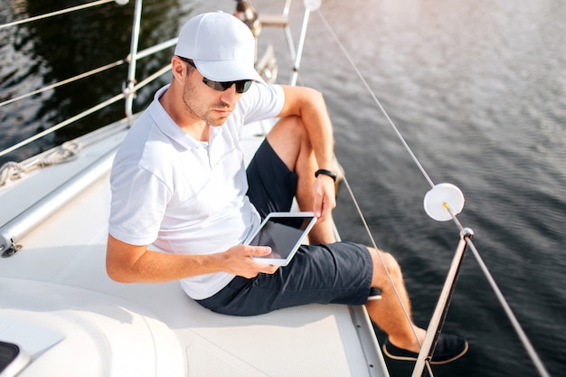 joven se sienta en el borde del tablero del yate y sostenga la tableta. El hombre lleva gafas de sol y gorra blanca con camisa. Él es serio y confiado. Sailor, descansa un poco.