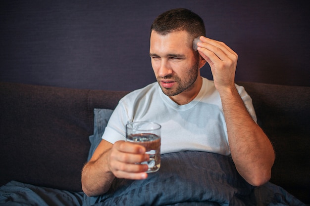 Joven se sienta en la cama y se encoge. El tiene dolor de cabeza. Guy mantiene una mano cerca de la cabeza y sostiene un vaso de agua con la otra. Está cubierto con una manta.