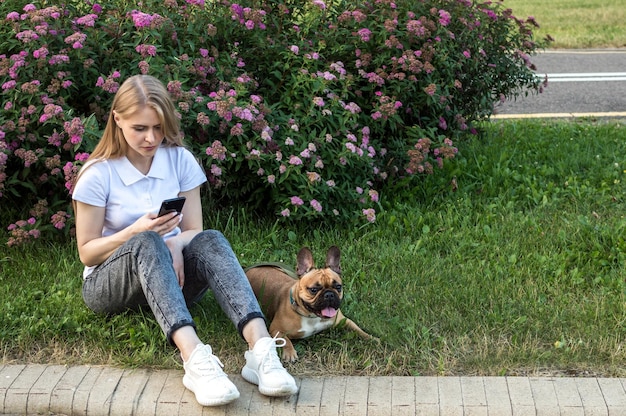 Una joven se sienta en el césped del parque y mira el teléfono. Un perro está sentado al lado.