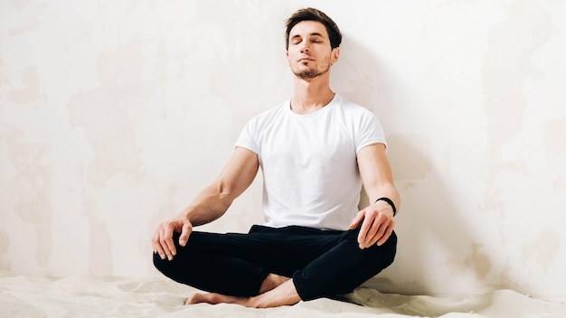 El joven se sienta en una pose meditativa sobre la arena contra un fondo de pared