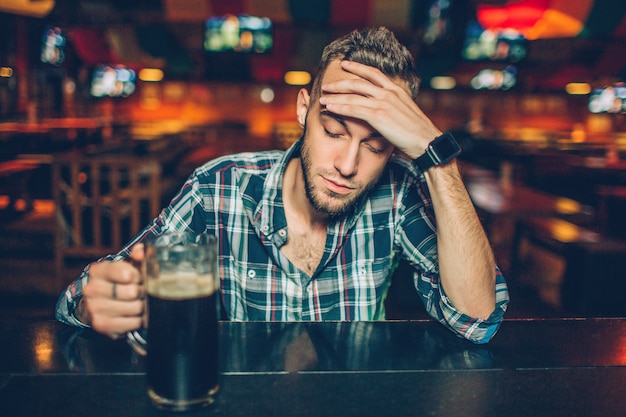 Un joven se sienta solo en la barra de bar en pub. Sostiene una jarra de cerveza oscura y mira hacia abajo. El joven tiene resaca.