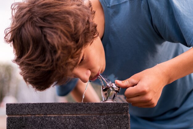 Joven siente sed, bebiendo agua de la fuente pública de la ciudad