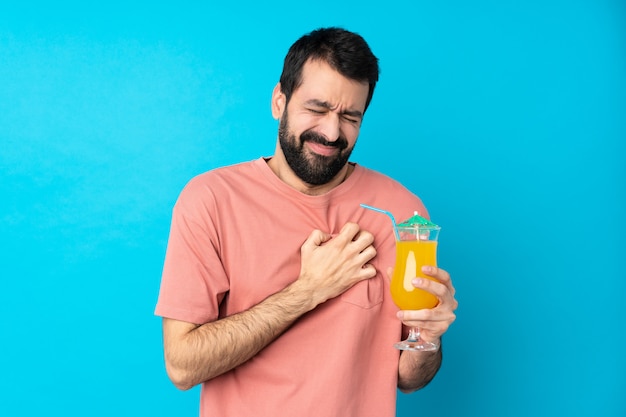 Joven sobre la celebración de un cóctel sobre la pared azul aislada que tiene un dolor en el corazón