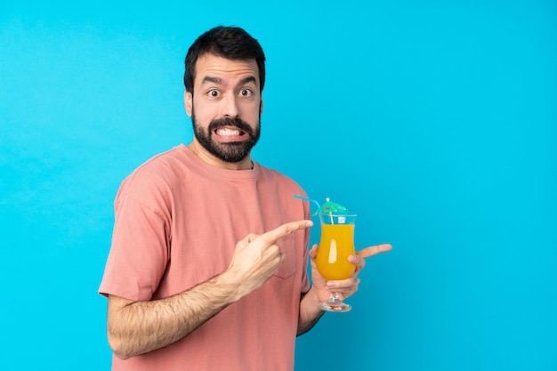 Joven sobre la celebración de un cóctel sobre la pared azul aislado asustado y apuntando hacia un lado