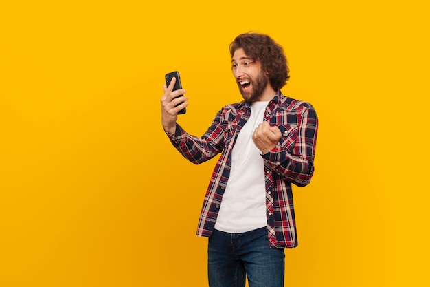 Un joven sobre un fondo amarillo mira un teléfono móvil y se regocija