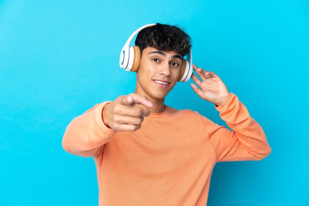 Foto joven sobre pared azul aislada escuchando música y apuntando hacia el frente