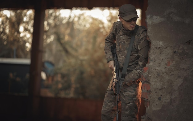 Un joven soldado ucraniano con experiencia en combate con un uniforme de camuflaje gris con gafas tácticas y una gorra con una ametralladora negra en las manos mira a la vuelta de la esquina en busca del enemigo