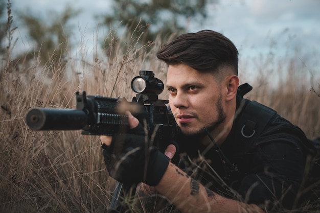Joven soldado en uniforme negro acostado y apuntando con un rifle de asalto
