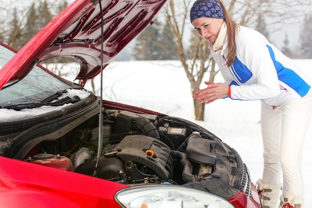 Una joven solitaria y preocupada con una chaqueta deportiva mirando el motor en el capó abierto del auto tratando de arreglar su auto rojo roto en un día nevado de invierno.