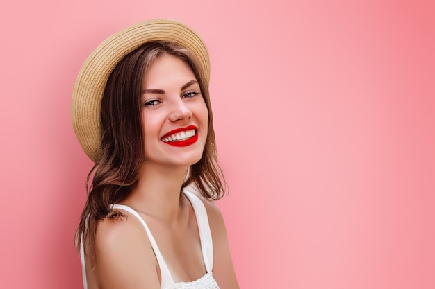 Una joven en sombrero de paja y lápiz labial rojo sonriendo en una pared de color rosa. Chica turista divirtiéndose. Niña feliz mirando a la cámara sonriendo y riendo espacio para texto