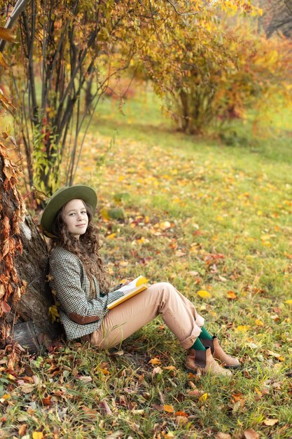 Una joven con sombrero y ropa de otoño se sienta en el césped con hojas doradas y un libro de lectura