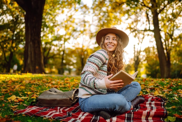 Una joven con sombrero se sienta en una manta en el parque de otoño y lee un libro Concepto de educación sobre la naturaleza