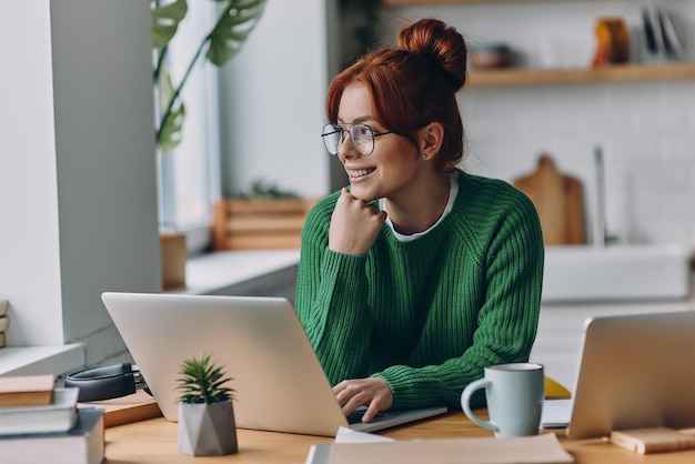 Una joven soñadora usando una computadora portátil y sonriendo mientras trabaja desde casa