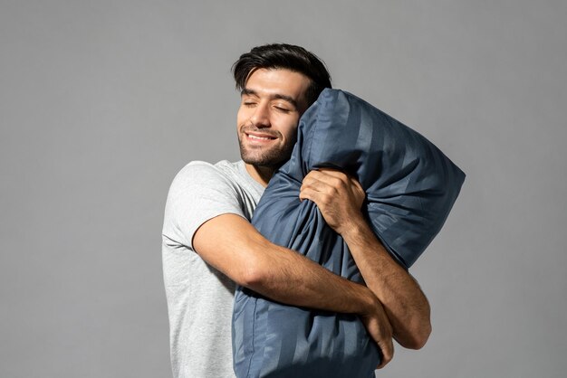 Un joven soñoliento sosteniendo una almohada cerrando los ojos soñando y sonriendo en un estudio aislado de fondo gris claro