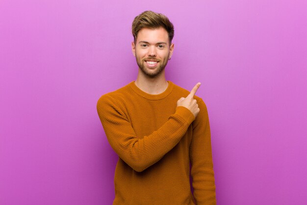 Joven sonriendo alegremente, sintiéndose feliz y apuntando hacia un lado y hacia arriba, mostrando el objeto en el espacio de la copia sobre la pared púrpura