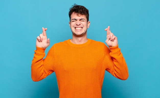 Joven sonriendo y cruzando ansiosamente ambos dedos, sintiéndose preocupado y deseando o esperando buena suerte